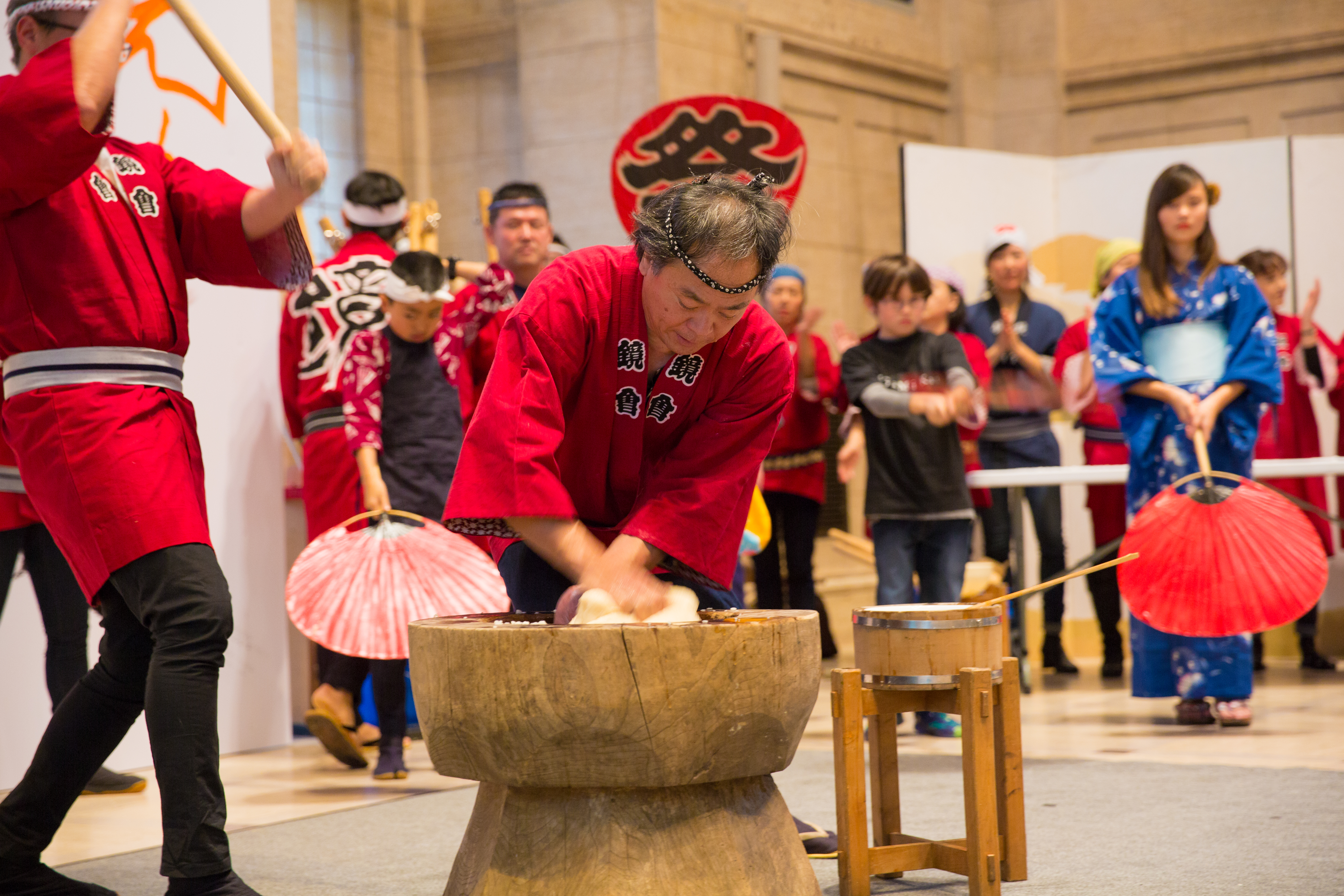 A man wearing a red tunic and black headband kneads mochi in a tall wooden bowl.