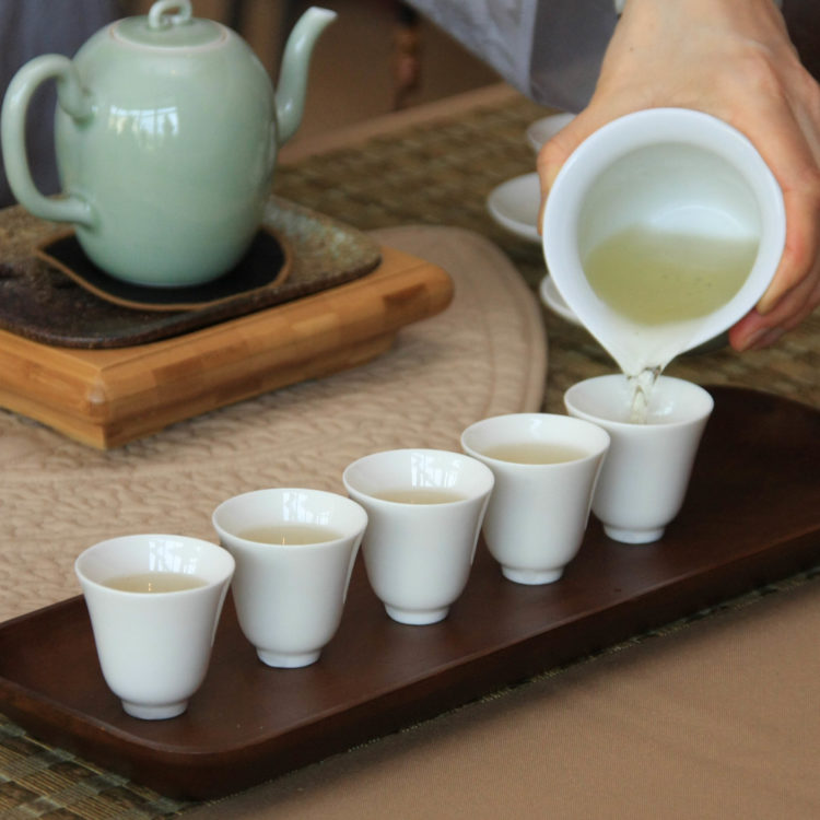 Tea is poured from a large white cup into five smaller white teacups.