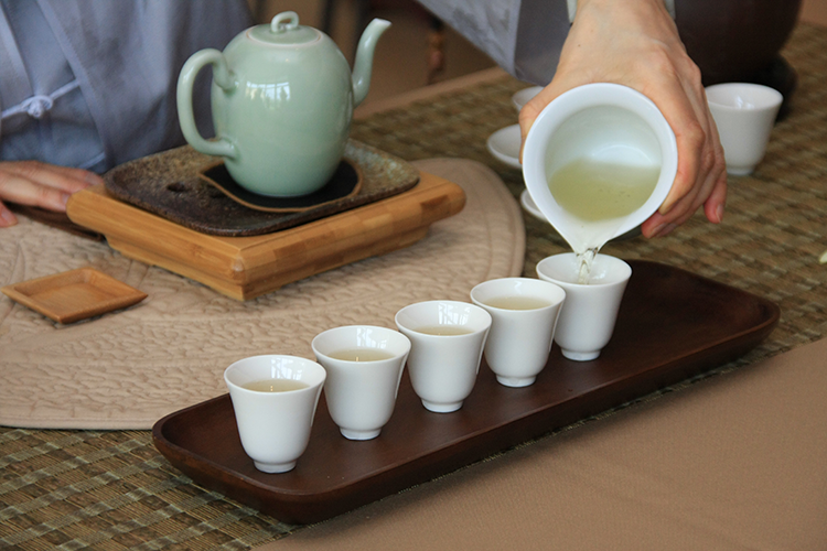 Tea is poured from a large white cup into five smaller white teacups.
