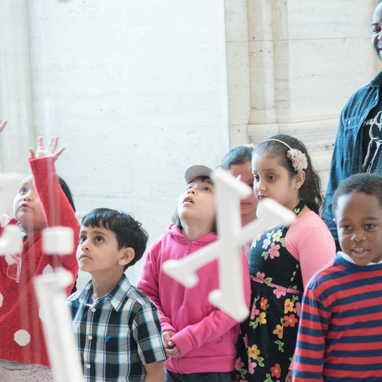 A group of small children and a chaperone observe an installation of white ceramic letters hanging from the ceiling.