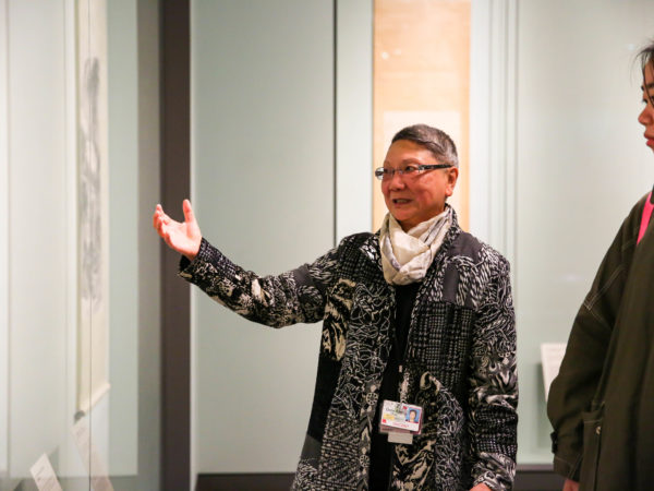 A docent gestures towards an artwork while a visitor looks on.