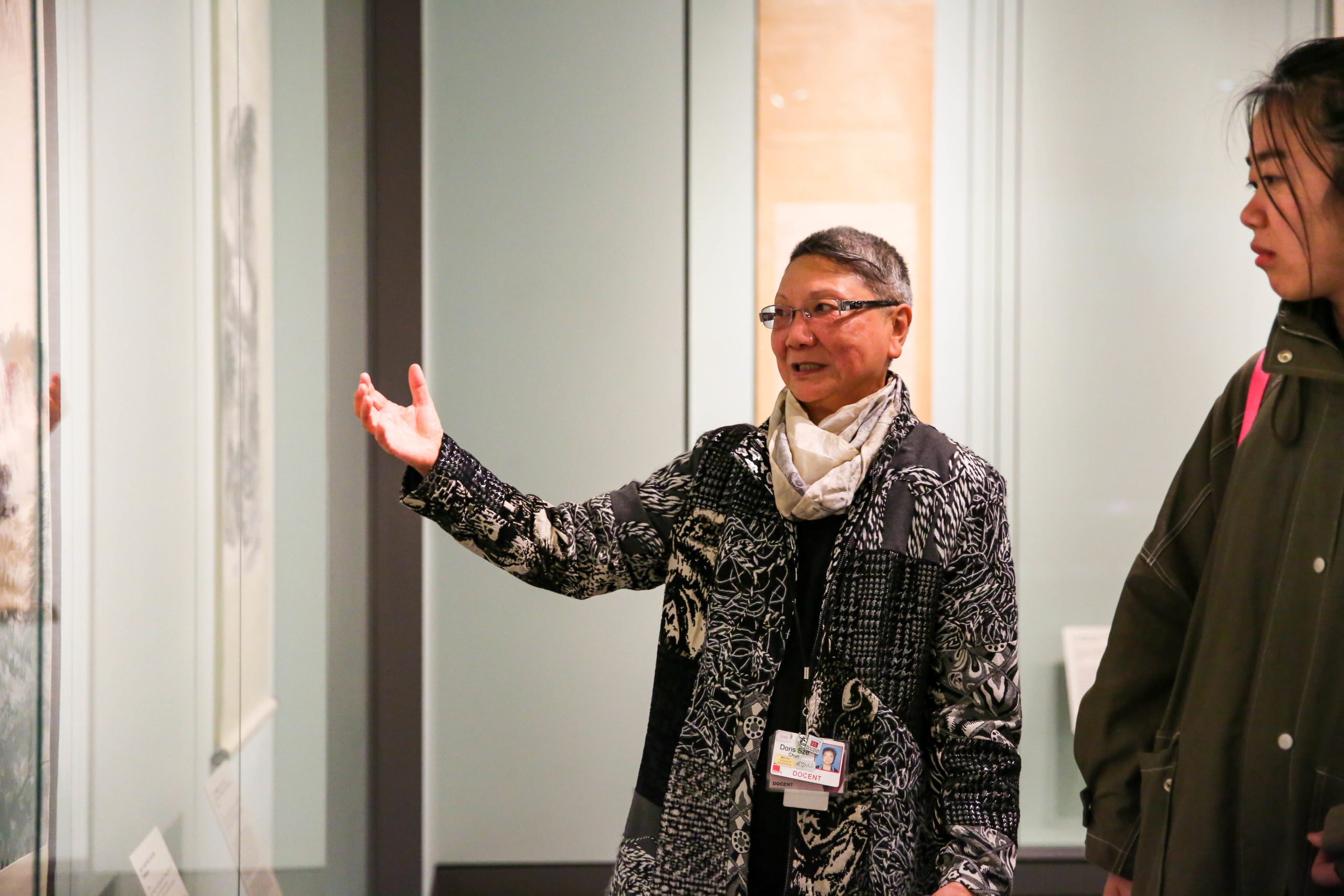 A docent gestures towards an artwork while a visitor looks on.