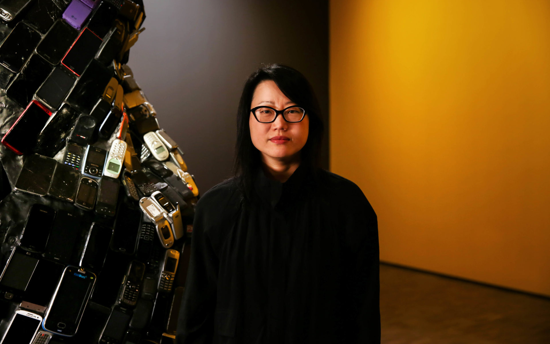Artist Jean Shin poses in the gallery with her installation, Pause, in front of a yellow wall and a sculpture covered in old cell phones.