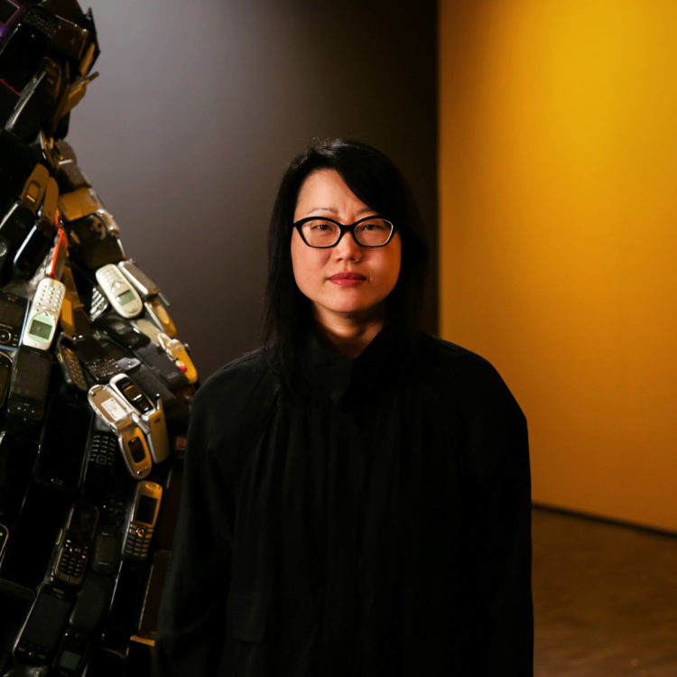 Artist Jean Shin poses in the gallery with her installation, Pause, in front of a yellow wall and a sculpture covered in old cell phones.