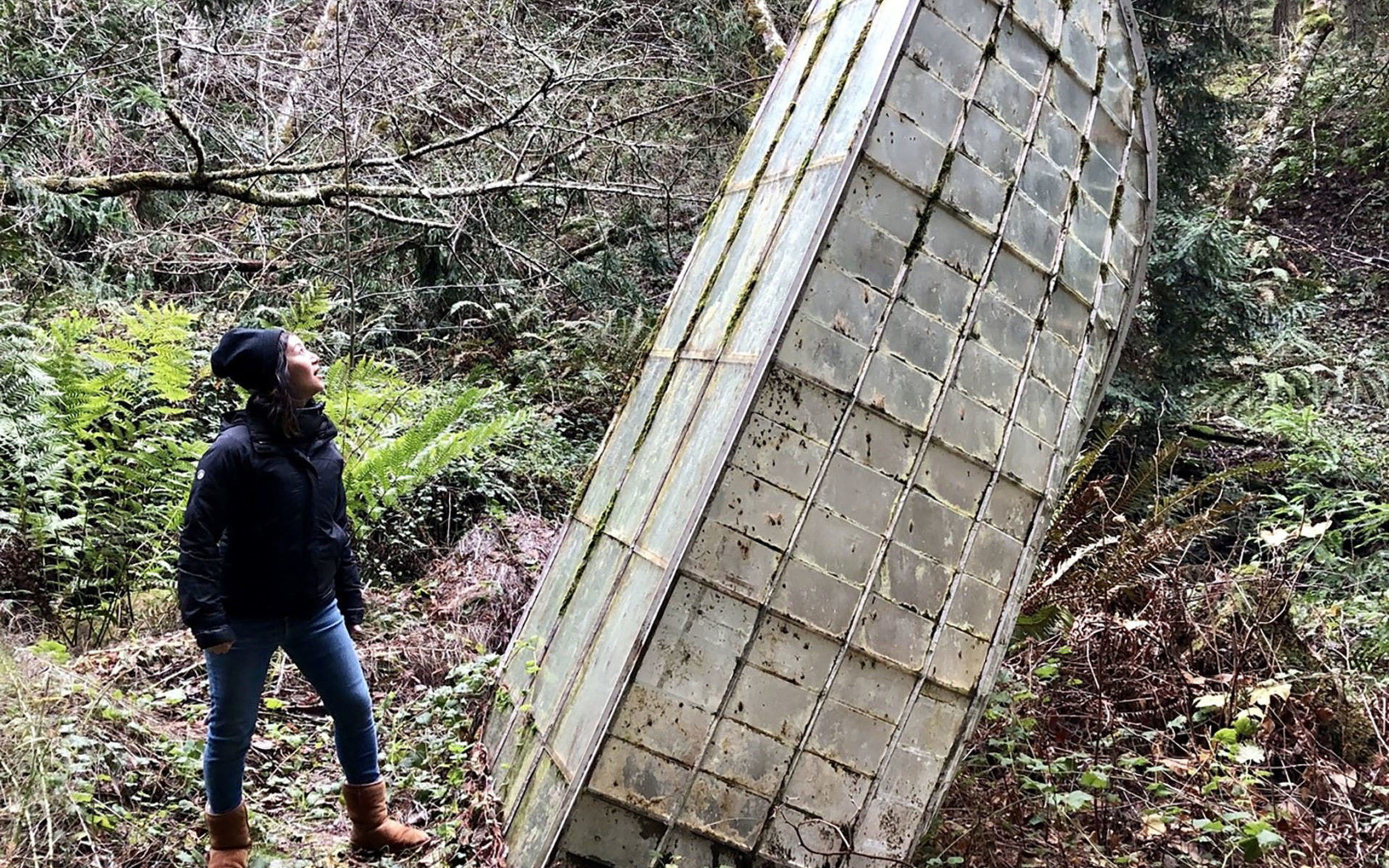 A woman looks at ruins of a tall glass structure in a forest.