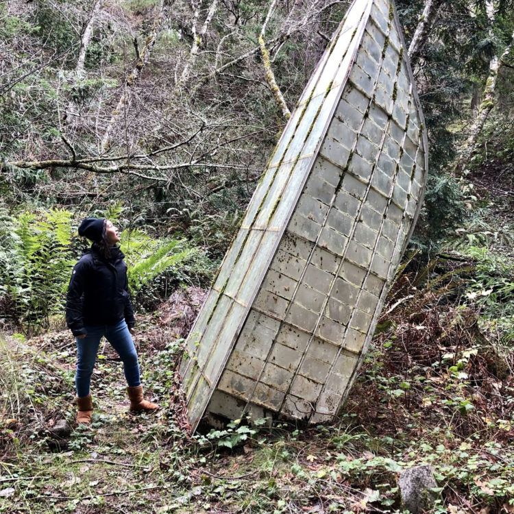 A woman looks at ruins of a tall glass structure in a forest.