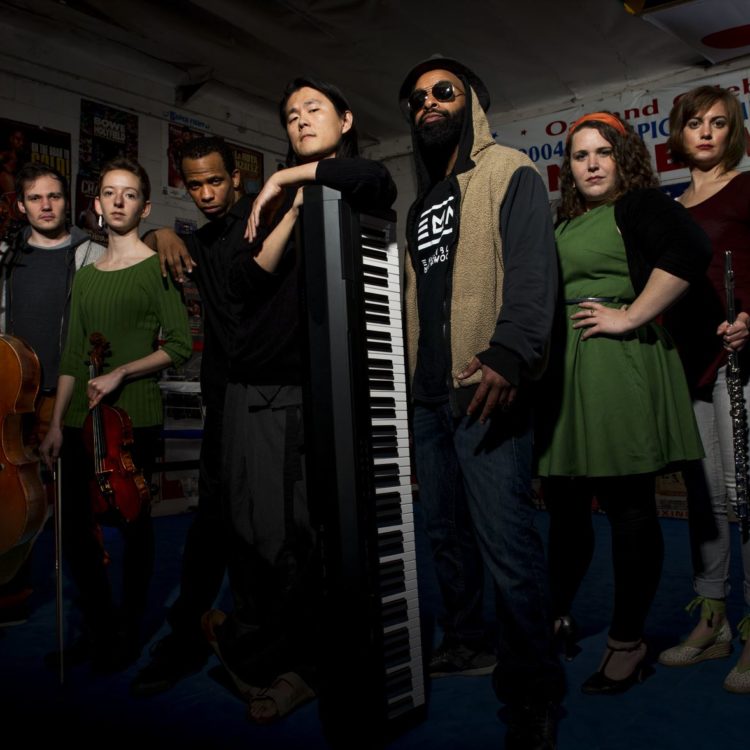 Nine musicians stand in a row holding their instruments in a dimly lit room.