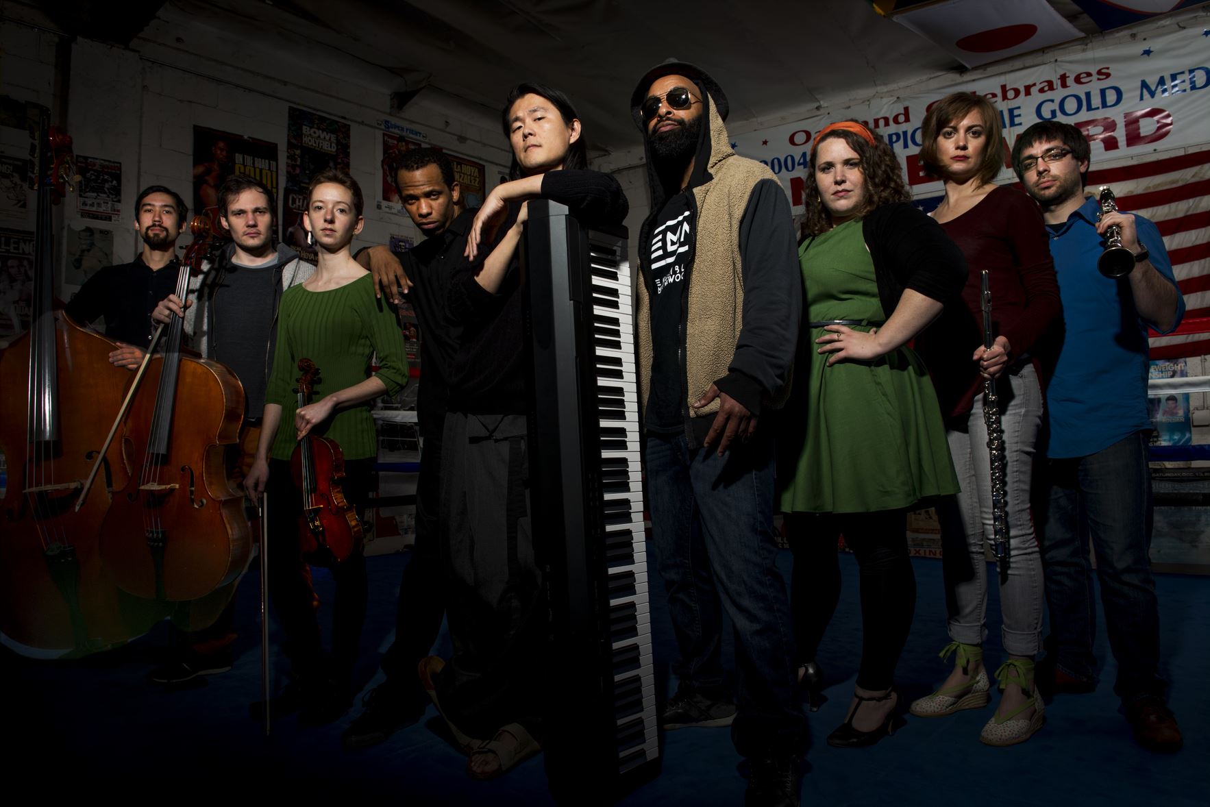 Nine musicians stand in a row holding their instruments in a dimly lit room.