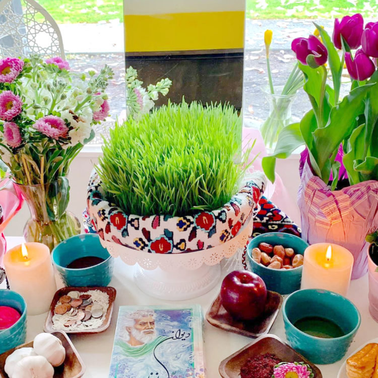 Table covered with colorful items, such as: a large, green wheatgrass plant; pink flowers; candles; shiny red apple; coins; and two small aqua bowls. 