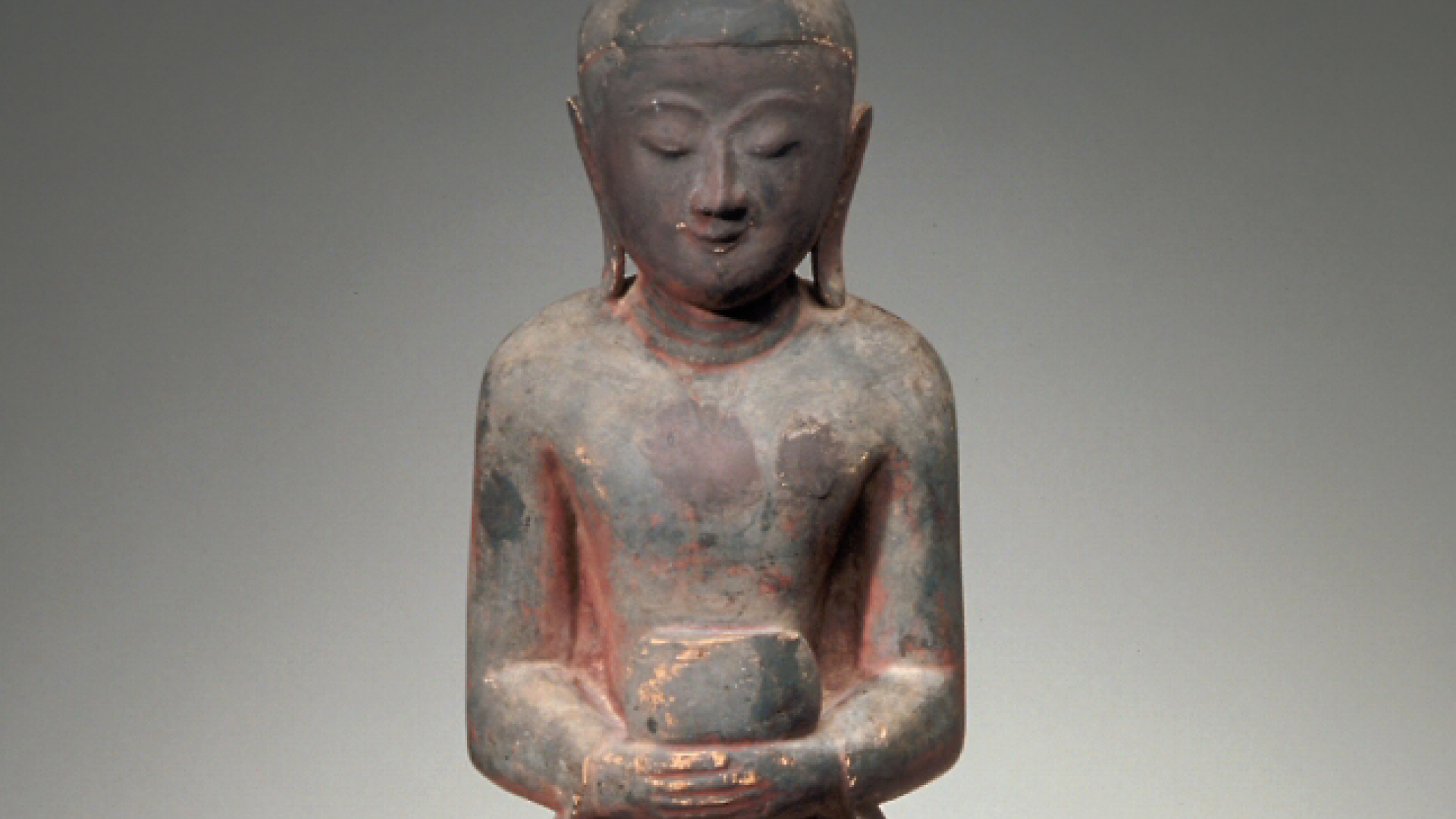 Wooden carved standing monk with downcast eyes and long earlobes, holding a bowl.