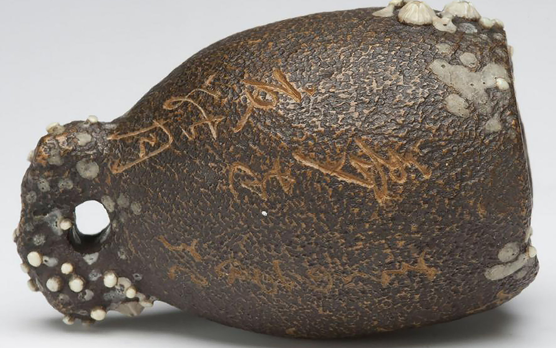 Thumb-sized carving of a brown, bell-shaped vessel covered with white barnacles and inscribed with cursive Japanese calligraphy in gold.