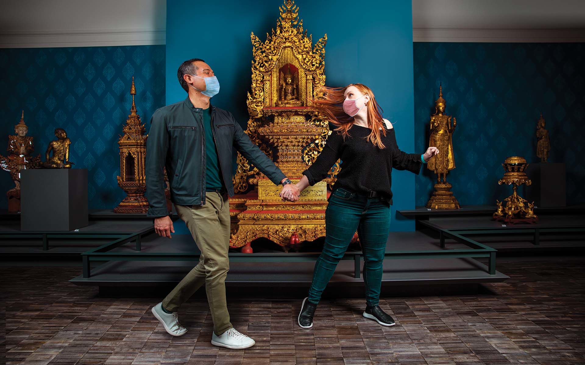 Two people wearing masks walk hand-in-hand through a gallery.