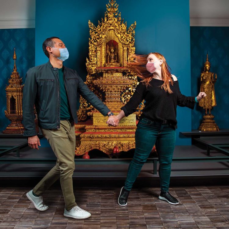 Two people wearing masks walk hand-in-hand through a gallery.