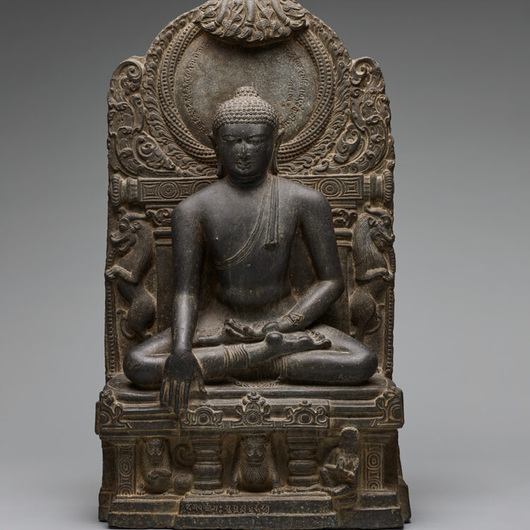 A 33-inch-tall sculpture finely carved in dark grey stone of a seated figure with legs folded. His left hand rests in his lap, palm facing the sky. His right hand reaches downward. Tightly coiled curls dot his head, including a lump at the top. A twisted garland of beads forms a circular halo behind and above his head.