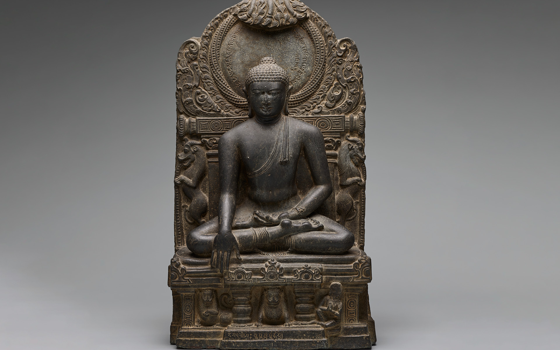 A 33-inch-tall sculpture finely carved in dark grey stone of a seated figure with legs folded. His left hand rests in his lap, palm facing the sky. His right hand reaches downward. Tightly coiled curls dot his head, including a lump at the top. A twisted garland of beads forms a circular halo behind and above his head.