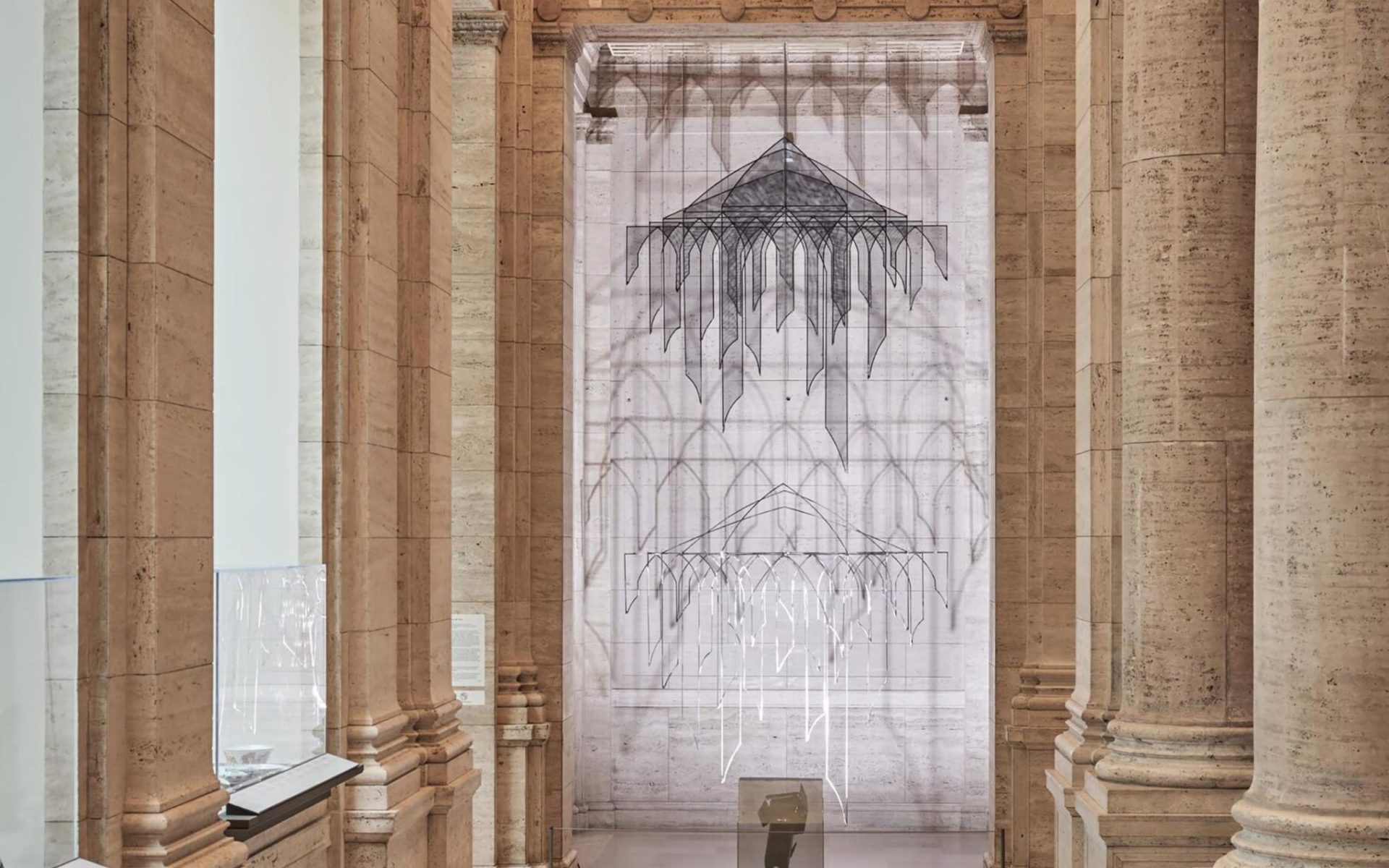 Chandelier-like hanging installation in silver and black wire and mesh forms of arches lit so that they cast shadows on the creamy marble walls of the gallery space.