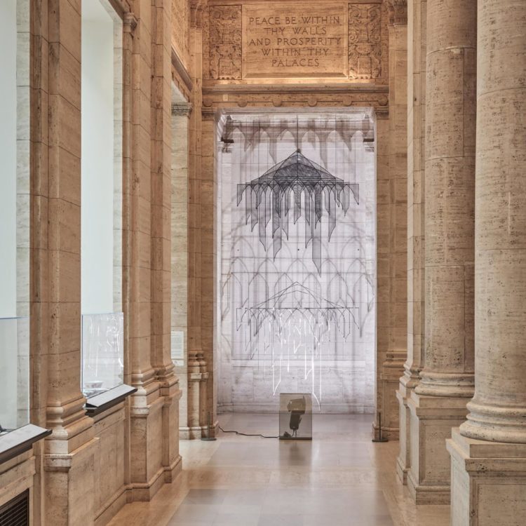 Chandelier-like hanging installation in silver and black wire and mesh forms of arches lit so that they cast shadows on the creamy marble walls of the gallery space.