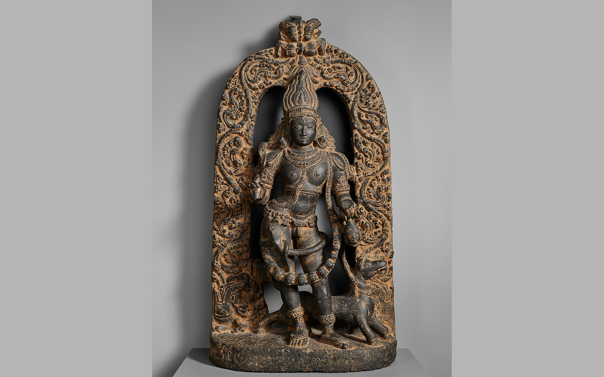 Sculpture of a crowned, bejeweled, and garlanded standing figure and a dog in dark stone with a brown substance rubbed into the indentations of the carving.