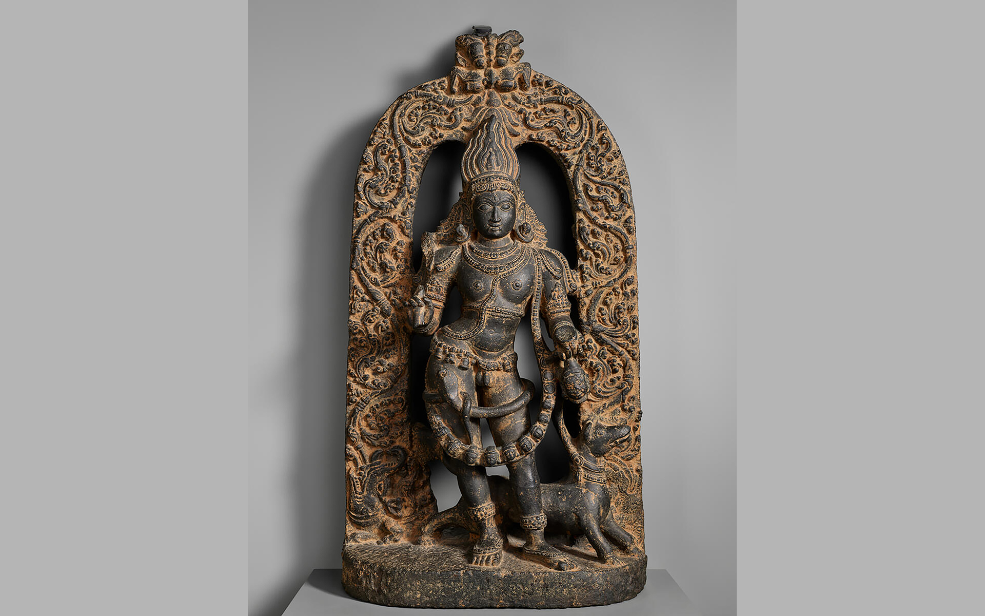 Sculpture of a crowned, bejeweled, and garlanded standing figure and a dog in dark stone with a brown substance rubbed into the indentations of the carving.