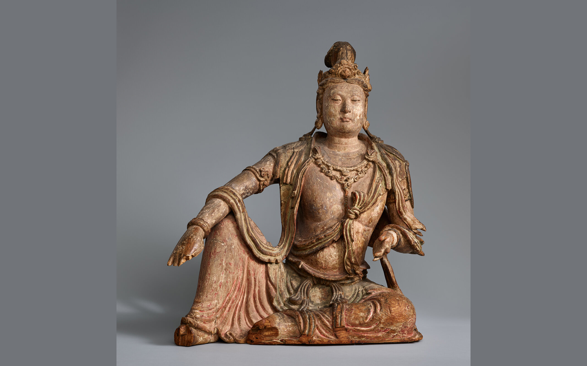 Larger than lifesize wooden crowned and bejeweled figure seated with right foot on the ground and right arm resting across the knee.