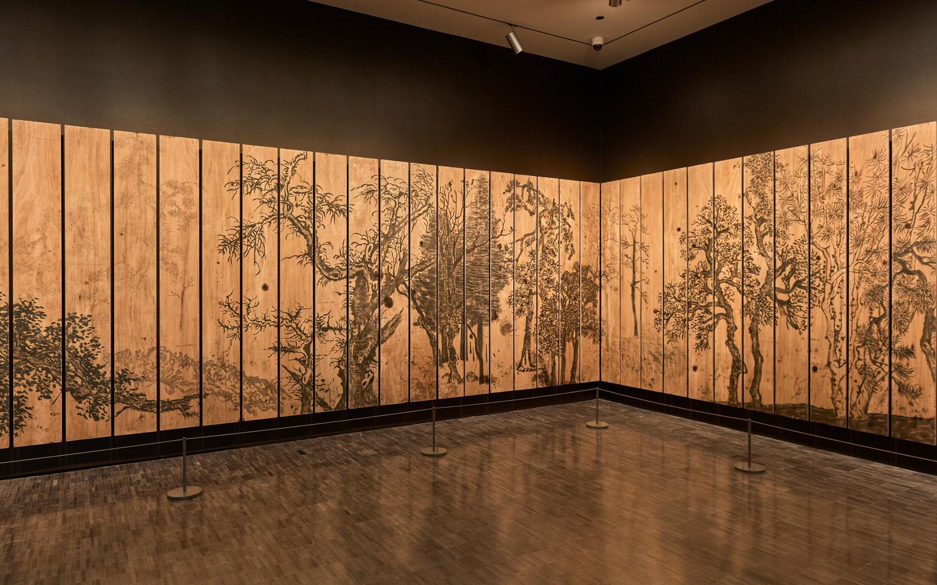 Corner of a gallery lined with vertical, warm brown-colored wooden panels painted with trees spanning across them.