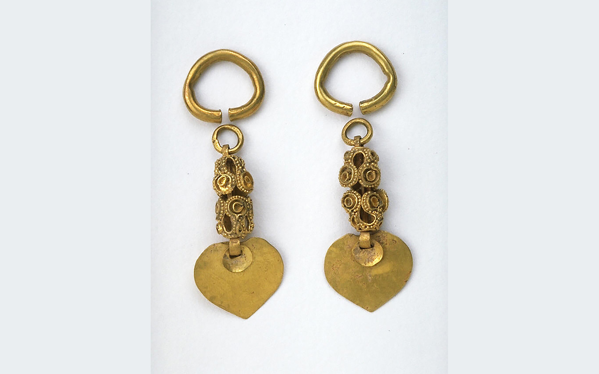 Pair of gold earrings with a large hoop at top, a chain of gold filigree and dots, and a flattened teardrop shape hanging at the bottom.