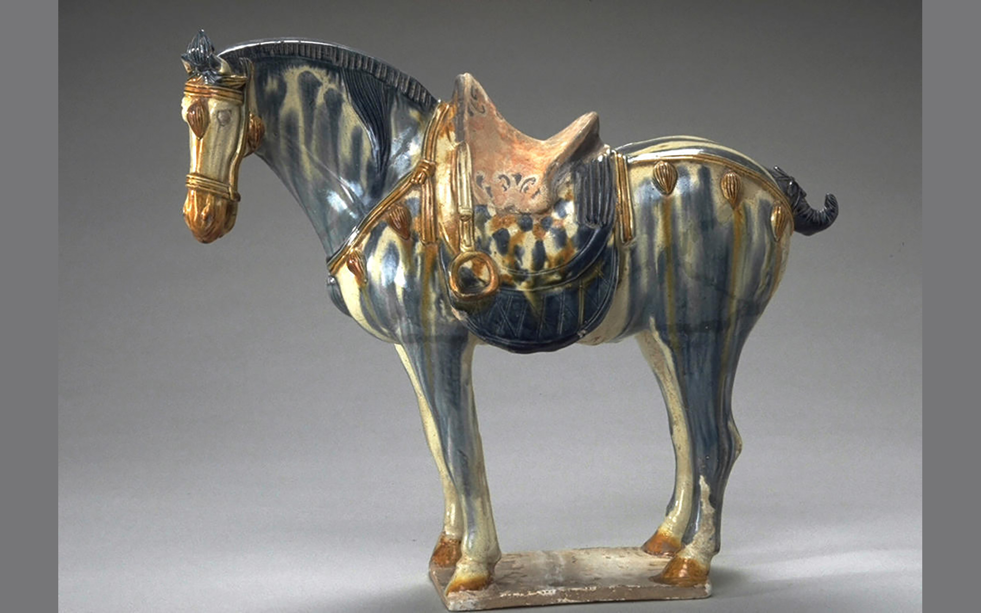 Ceramic horse with bridle and saddle glazed in blue, cream, and brown that stands over one foot tall.