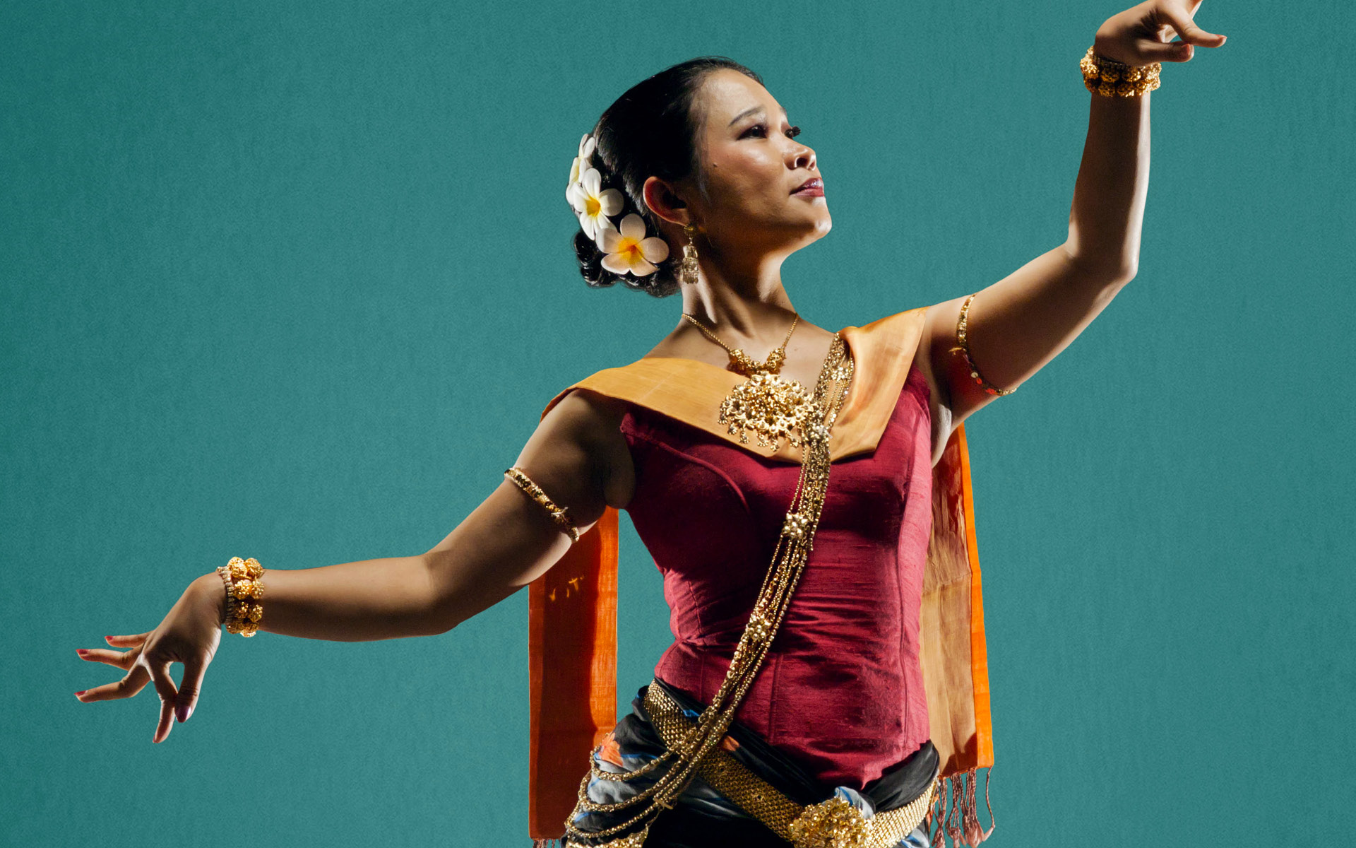 Single female dancer standing at an angle with both arms in the air, one lifted up past her shoulder, the other behind her and lower. Her left leg is bent and the right leg extended behind her. She is wearing a red sleeveless top with a gold neck, dark pants and her hair in a bun held with flowers.