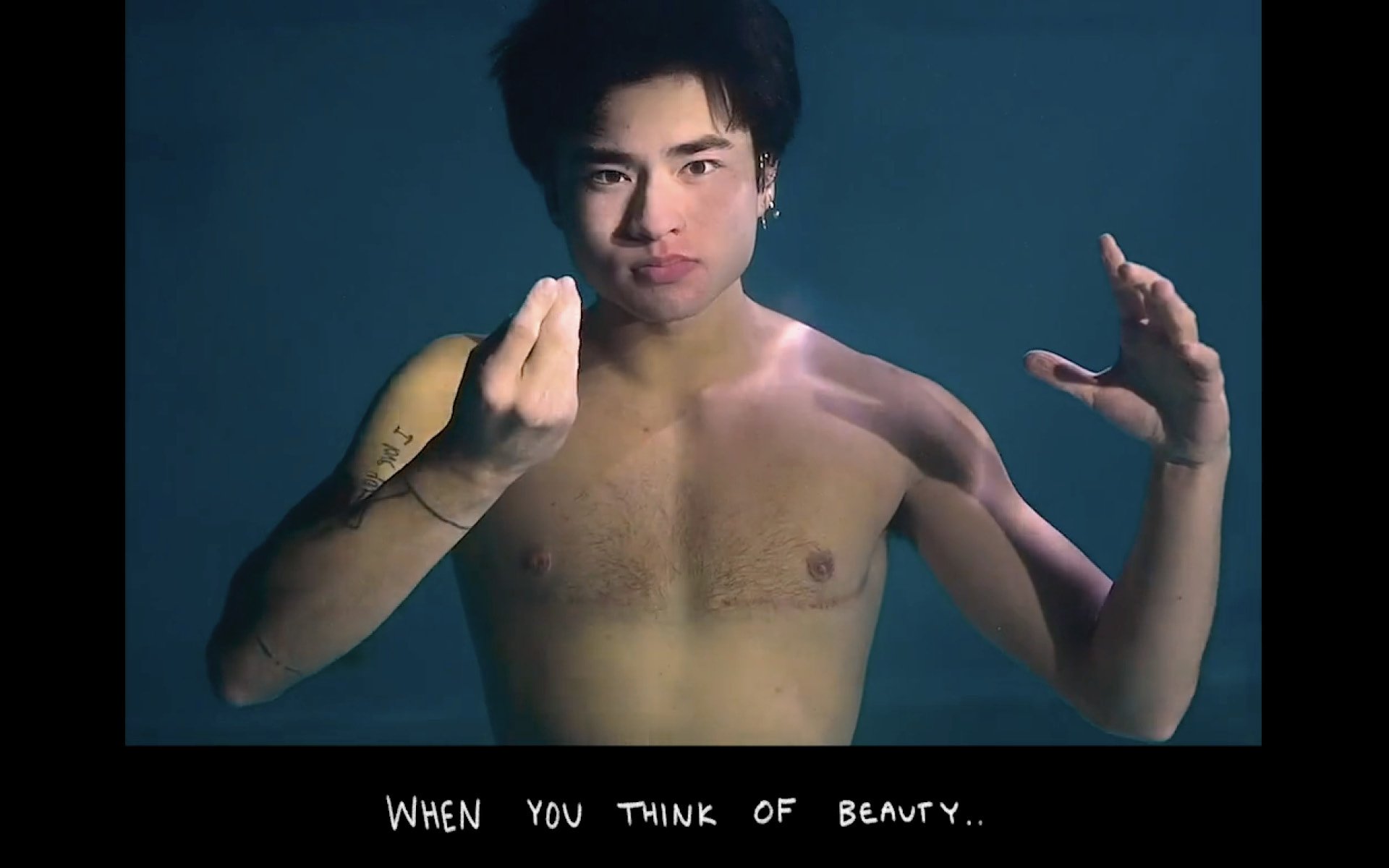 Chellaman, who is an Asian American Deaf transman poses underwater, his black hair floating above his head, and his hand making an ASL sign in front of his chest, where is top surgery scars can be seen. Text reads: “When you think of beauty…”