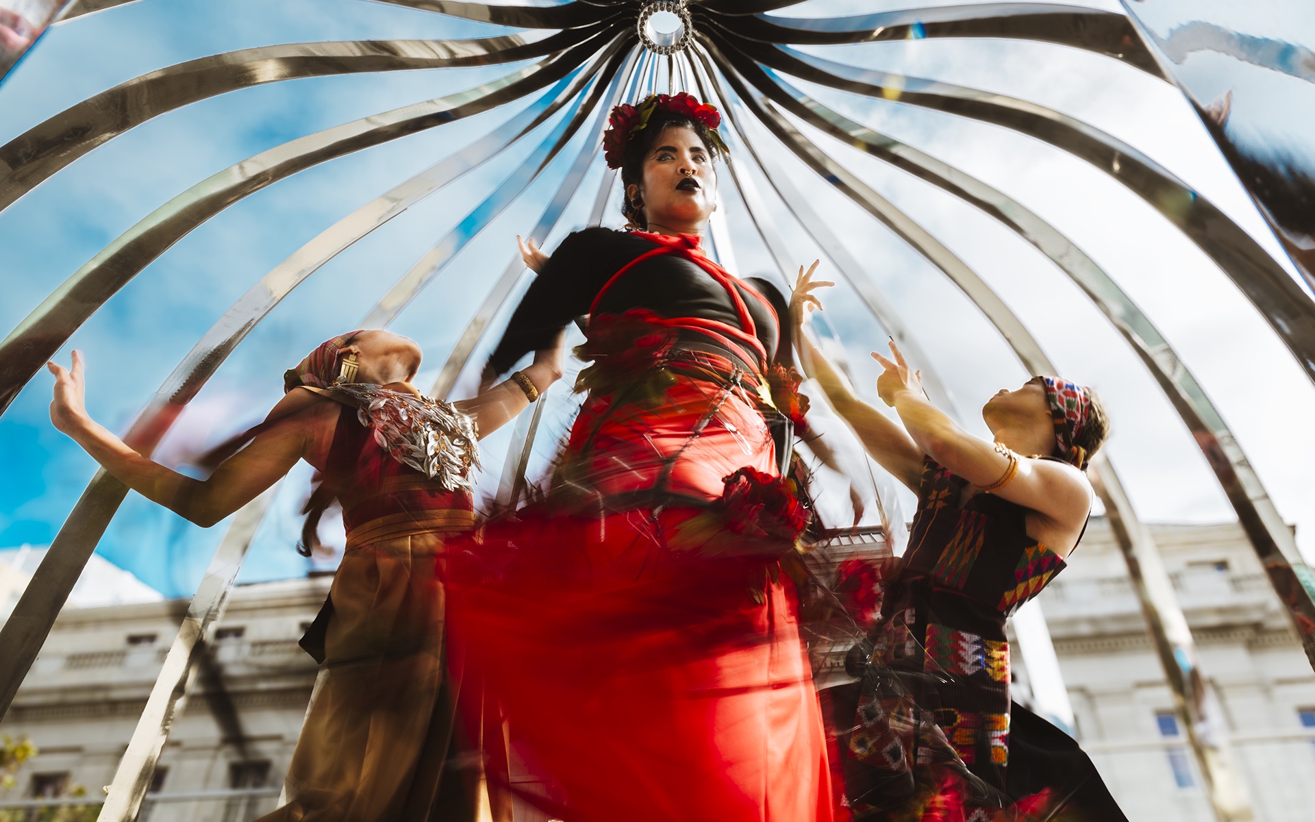 Three dancers photographed from below underneath a metal structure through which we can see blue sky. The center dancer wearing a long red skirt moves creating a blur. Two dancers in woven textiles frame her; they look away from the viewer and up to the sky.